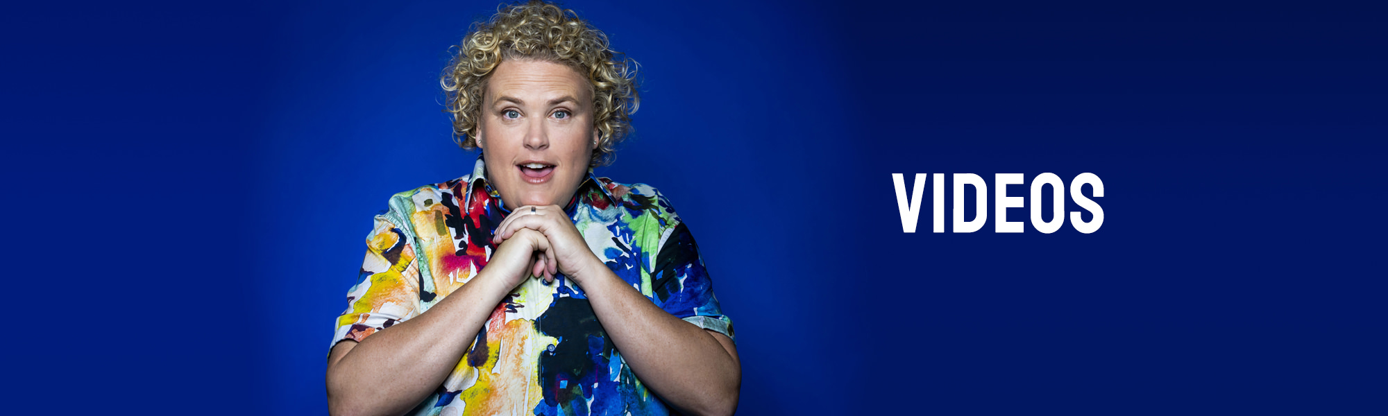 Comedian Fortune Feimster in front of a dark blue background with her hands clasped beneath her chin.
